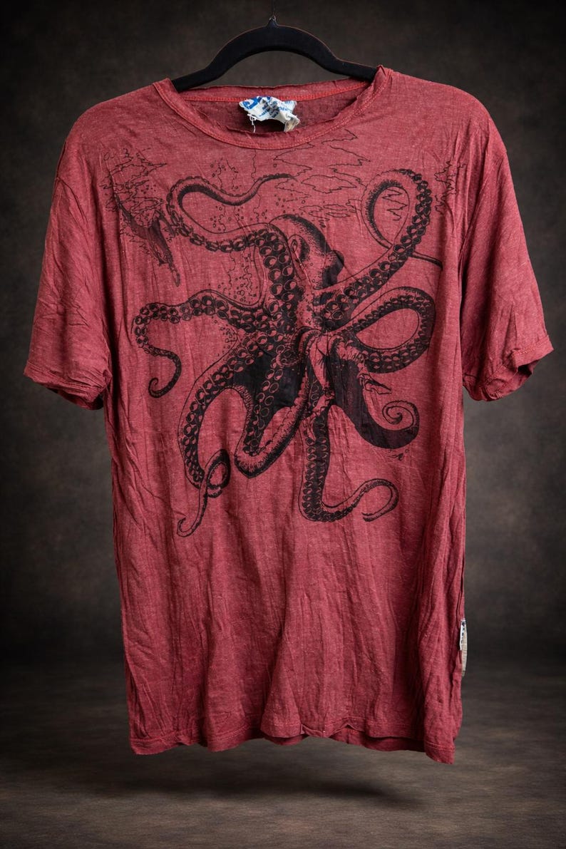 May include: A short-sleeved, crew-neck t-shirt in a faded red color. The front features a large, detailed black ink-style illustration of an octopus with its tentacles spread across the chest area. The fabric has a slightly textured, worn appearance.