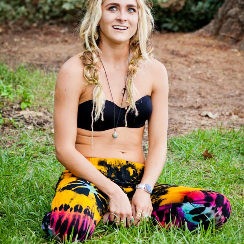 May include: A woman with blonde hair in braids sits on the grass wearing a black bandeau top and colorful tie-dye pants.