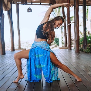 May include: A woman wearing a black crop top and blue patterned pants with a side slit. She is stretching in a yoga pose on a wooden deck.