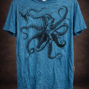 May include: A blue short-sleeve t-shirt featuring a large black graphic print of an octopus with detailed tentacles and suction cups. The shirt has a crew neckline and a small rectangular brand tag attached to the lower right side seam.