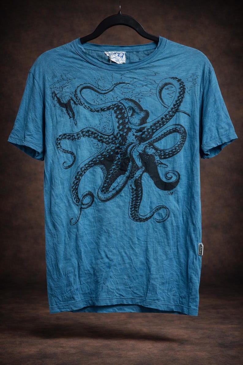 May include: A blue short-sleeve t-shirt featuring a large black graphic print of an octopus with detailed tentacles and suction cups. The shirt has a crew neckline and a small rectangular brand tag attached to the lower right side seam.