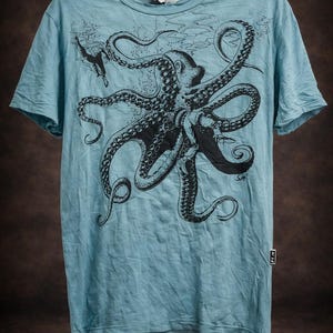 May include: A light blue short-sleeve t-shirt featuring a large black graphic print of an octopus with long, detailed tentacles. The design includes a small diver figure near the top left. A small white rectangular tag is visible on the side seam.