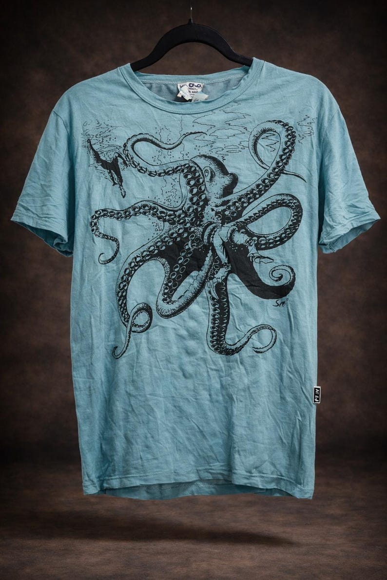 May include: A light blue short-sleeve t-shirt featuring a large black graphic print of an octopus with long, detailed tentacles. The design includes a small diver figure near the top left. A small white rectangular tag is visible on the side seam.