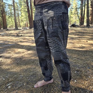 May include: Pants featuring a patchwork design with various black and gray geometric patterns, including houndstooth and hexagonal motifs. The pants have a high waistband, a button closure on the side pocket, and a tapered leg silhouette.
