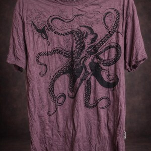 May include: A mauve short-sleeved t-shirt featuring a large black graphic print of an octopus with extended tentacles. The shirt has a crew neckline, a visible brand tag at the inner collar, and a small rectangular brand label on the lower side seam.