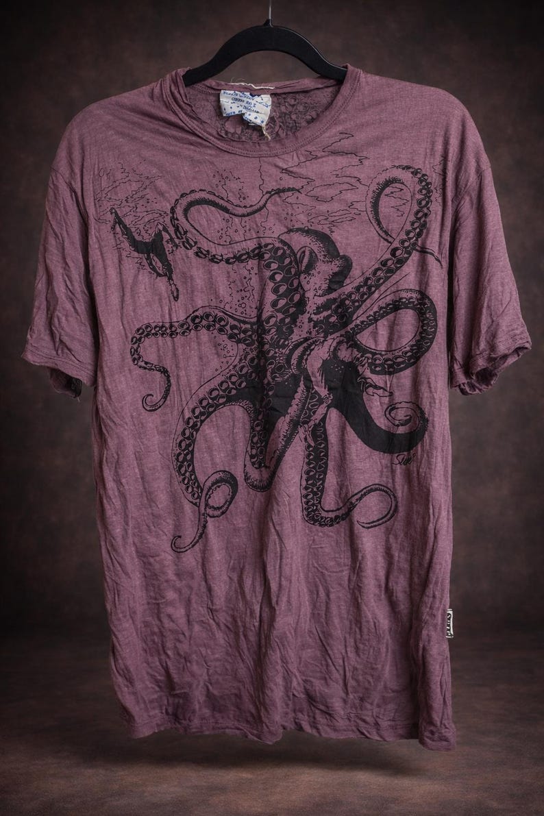 May include: A mauve short-sleeved t-shirt featuring a large black graphic print of an octopus with extended tentacles. The shirt has a crew neckline, a visible brand tag at the inner collar, and a small rectangular brand label on the lower side seam.