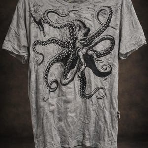 May include: A heather gray short-sleeve t-shirt featuring a large black ink-style illustration of an octopus with long tentacles. The graphic includes a small diver figure and subtle water textures. A small white tag is visible on the side seam.
