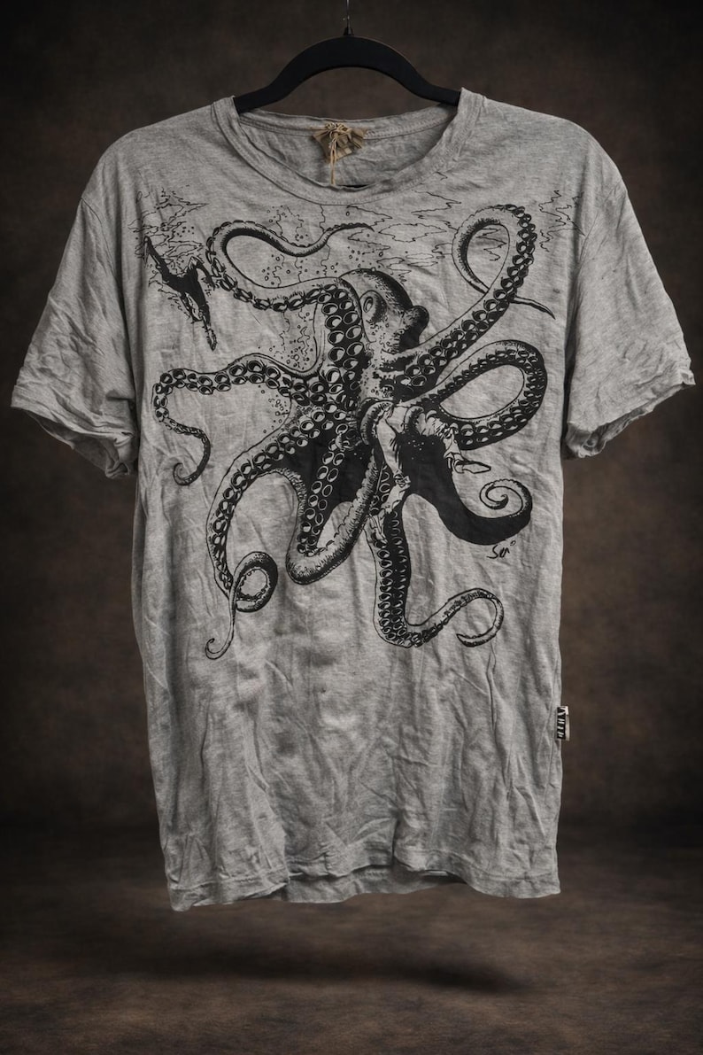 May include: A heather gray short-sleeve t-shirt featuring a large black ink-style illustration of an octopus with long tentacles. The graphic includes a small diver figure and subtle water textures. A small white tag is visible on the side seam.