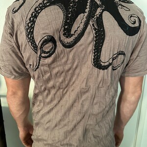 May include: A taupe-colored short-sleeve t-shirt featuring a large, black graphic print of an octopus on the back. The octopus design covers the upper back area, showcasing detailed tentacles with visible suction cups extending across the shoulders.