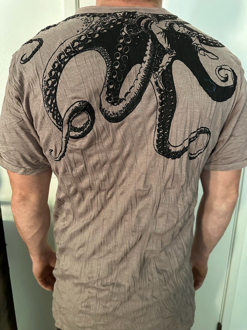 May include: A taupe-colored short-sleeve t-shirt featuring a large, black graphic print of an octopus on the back. The octopus design covers the upper back area, showcasing detailed tentacles with visible suction cups extending across the shoulders.
