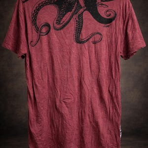 May include: A short-sleeved t-shirt in a heathered maroon color featuring a large black graphic print of an octopus across the upper back and shoulder area. The design includes detailed tentacles and a small signature text near the right side.