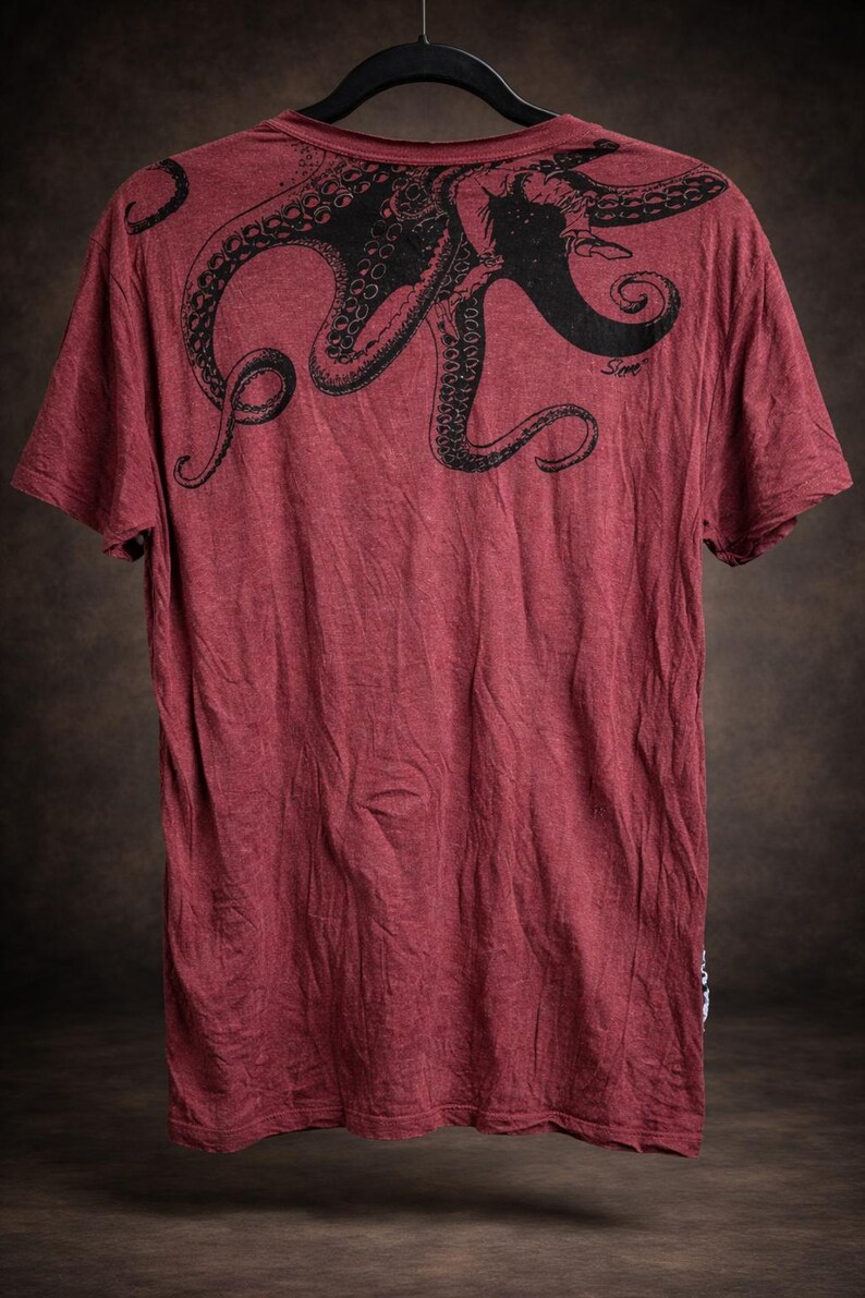 May include: A short-sleeved t-shirt in a heathered maroon color featuring a large black graphic print of an octopus across the upper back and shoulder area. The design includes detailed tentacles and a small signature text near the right side.