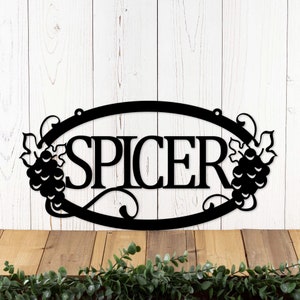 May include: Black metal sign with the name "SPICER" in a decorative font, surrounded by grapevine designs. The sign is oval shaped and has a rustic look.