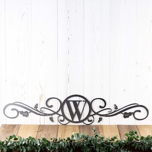 May include: Black metal monogram with a decorative scroll design and the letter "W" in the center. The monogram is mounted on a white wood background.