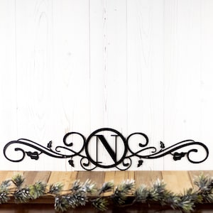 May include: Black metal monogram with a decorative scroll design and the letter "N" in a circle. The monogram is mounted on a white wood background.