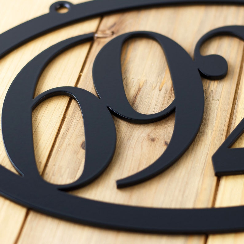 Close up of matte black powder coat from our horizontal oval 4 digit metal house number sign. Placed on a wood plank.