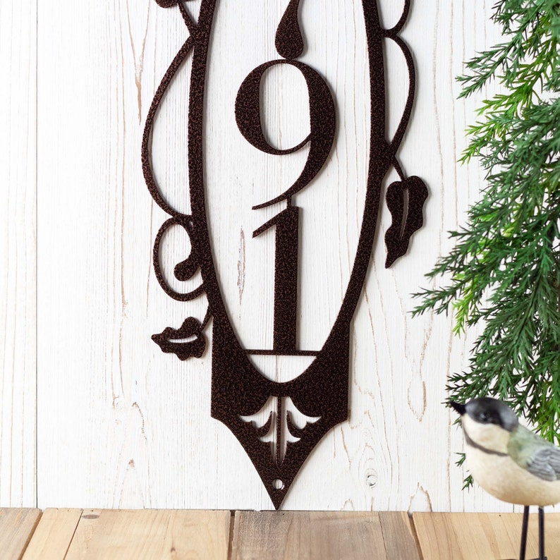Close up of fleur de lis and mounting hole on our vertical 3 digit metal house number plaque, in copper vein powder coat. Placed against a white wood wall.