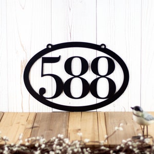 Horizontal oval 3 digit metal house number sign, in matte black powder coat. Placed against a white wood wall.