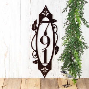 3 digit vertical metal house number plaque with vines and fleur de lis, in copper vein powder coat. Placed against a white wood wall.