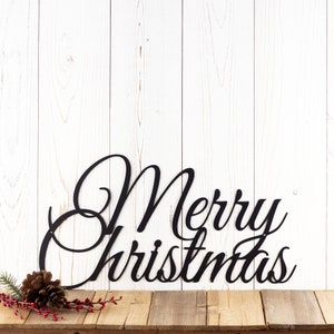 Merry Christmas script metal wall art, in matte black powder coat. Placed against a white wood wall.