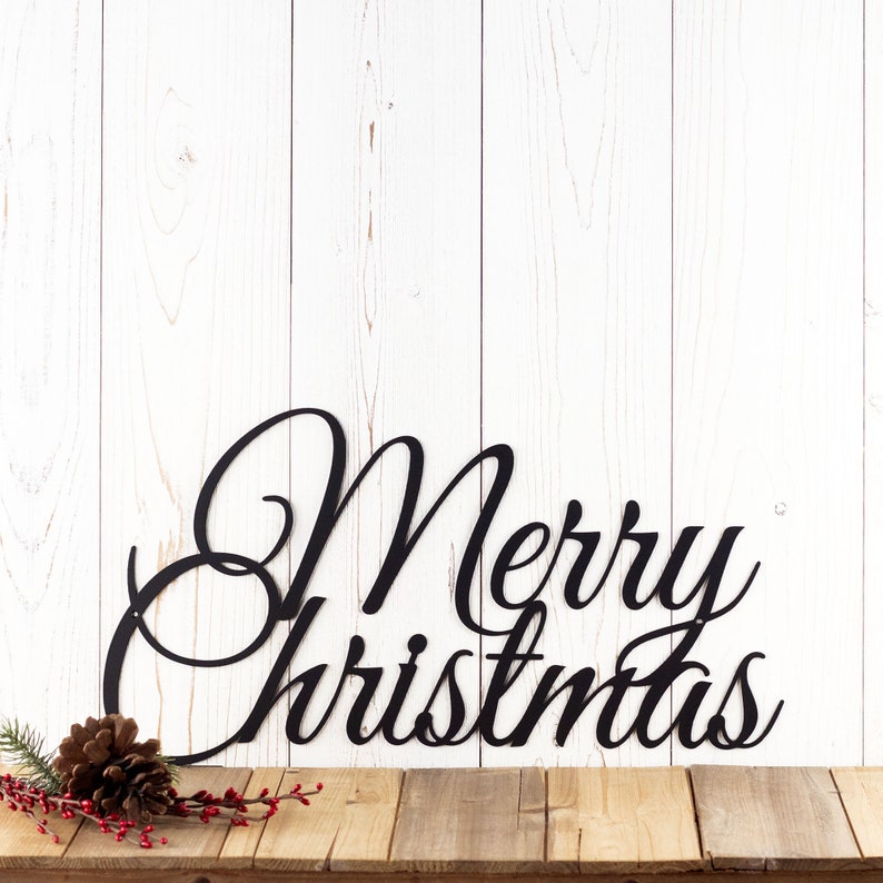 Merry Christmas script metal wall art, in matte black powder coat. Placed against a white wood wall.