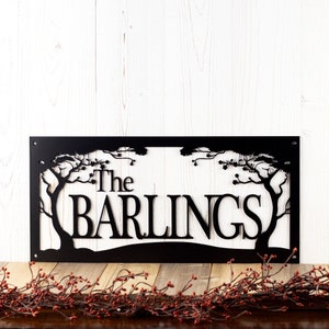 Rectangular metal family name plaque with cherry trees, in matte black powder coat. Placed against a white wood wall.