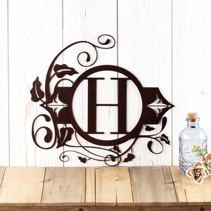 May include: A metal monogram with a decorative floral design. The letter "H" is in a circle in the center of the design. The monogram is painted in a dark brown color.
