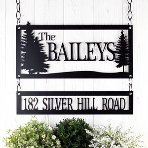 May include: Black metal sign with two pine trees on either side. The sign reads "The Baileys" on the top line and "182 Silver Hill Road" on the bottom line.