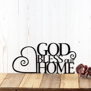 God Bless our Home metal wall art with heart, in matte black powder coat. Placed against a white wood wall.