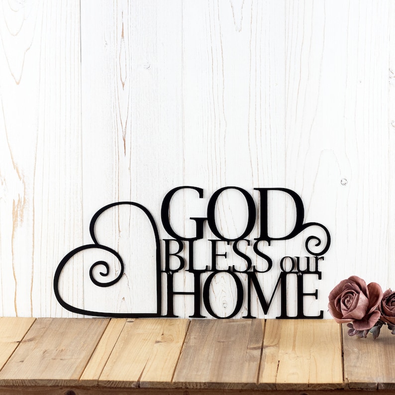 God Bless our Home metal wall art with heart, in matte black powder coat. Placed against a white wood wall.