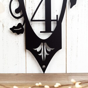 Close up of fleur de lis and mounting hole on our vertical 3 digit metal house number sign, in matte black powder coat. Placed against a white wood wall.