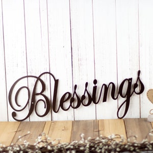 May include: A metal wall decor sign with the word "Blessings" in a decorative script font. The sign is black and is mounted on a white wood background.