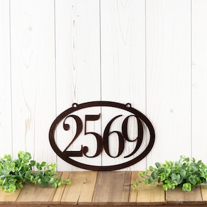 Horizontal oval 4 digit metal house number sign, in copper vein powder coat. Placed against a white wood wall.