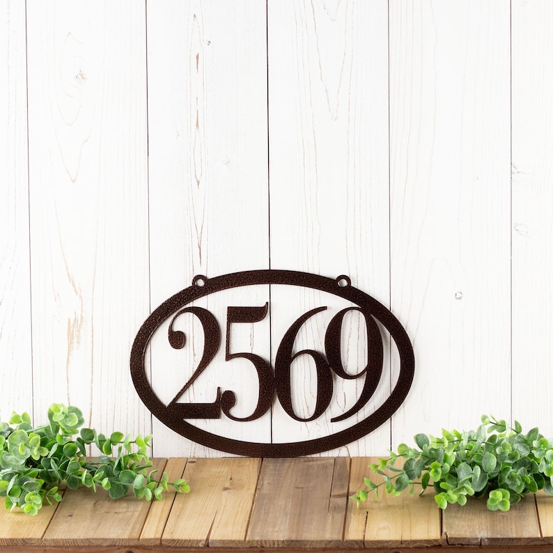 Horizontal oval 4 digit metal house number sign, in copper vein powder coat. Placed against a white wood wall.