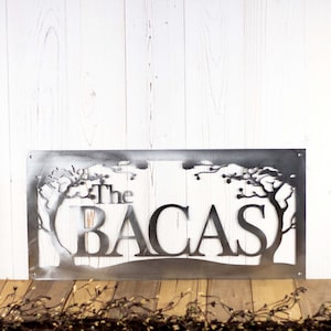 Rectangular metal family name sign with cherry trees, in raw steel. Placed against a white wood wall.