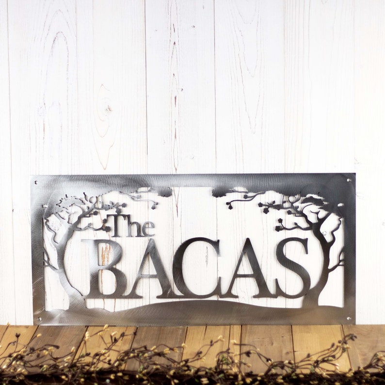 Rectangular metal family name sign with cherry trees, in raw steel. Placed against a white wood wall.