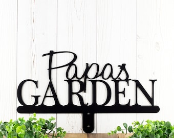 May include: Black metal sign with the words "Papa's Garden" in a decorative font. The sign is mounted on a white wooden background.