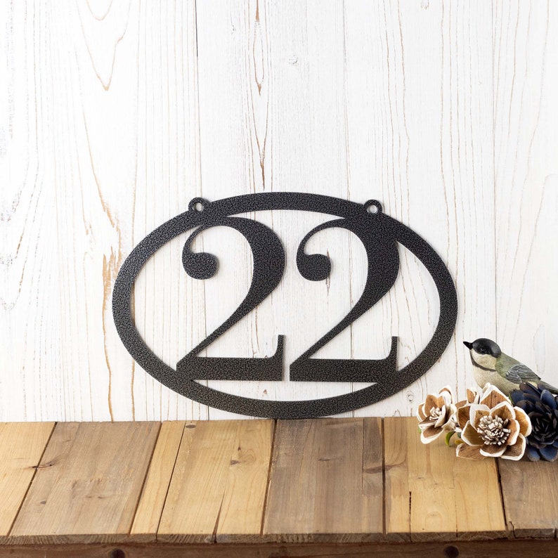 Horizontal oval 2 digit metal house number sign, in silver vein powder coat. Placed against a wood wall.