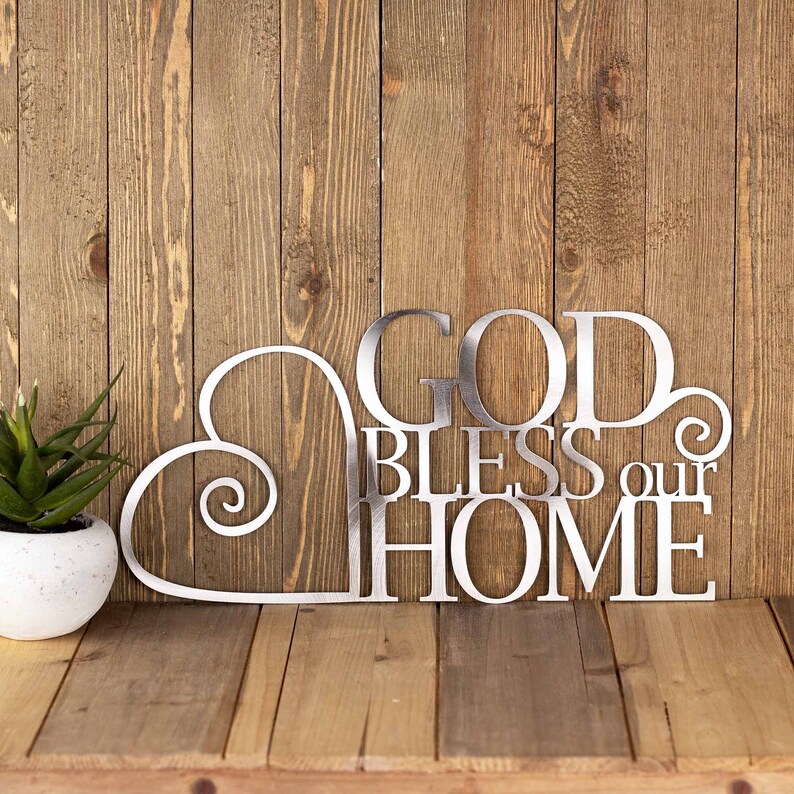 God Bless our Home metal plaque with a heart, in raw steel. Placed against a wood wall.
