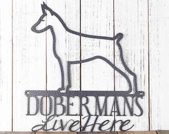 May include: Black metal silhouette of a Doberman Pinscher dog with the text "Dobermans Live Here" below. The silhouette is mounted on a white wooden background.