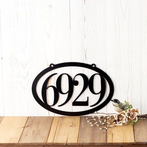 4 Digit oval metal house number sign with 2 mounting holes, in matte black powder coat. Placed against a white wooden wall with white garland.