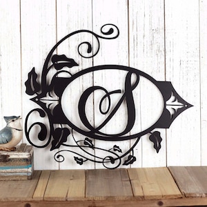May include: Black metal monogram with a decorative floral design and the letter "S" in a circle. The monogram is mounted on a white wood background.