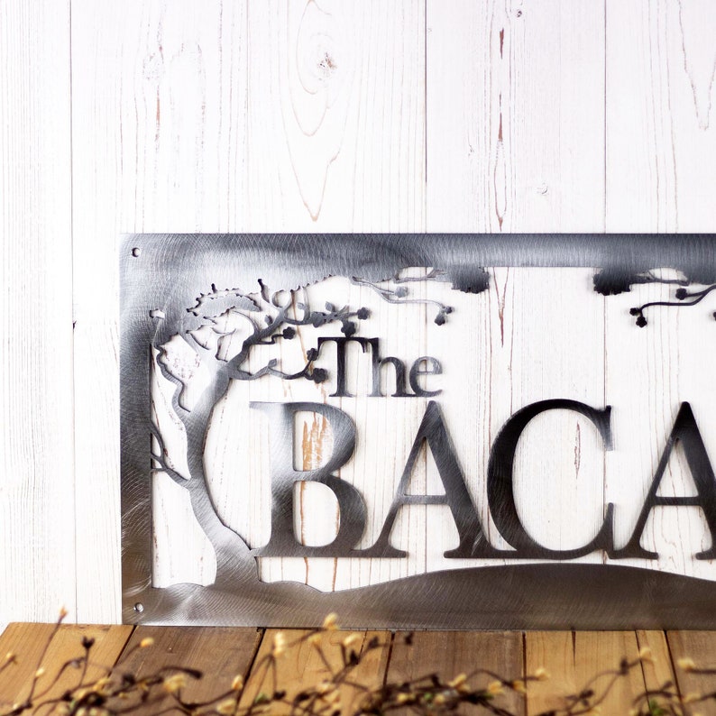 Close up of metal family last name sign with cherry trees, in raw steel. Placed on a white wood plank.