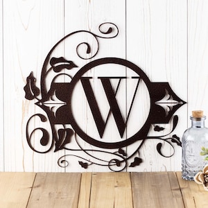 May include: A metal monogram sign with a decorative floral design. The letter "W" is in a circle in the center of the sign. The sign is brown and has a rustic finish.