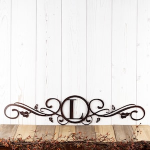 May include: A metal wall decor with a scrolled design and a circle in the center with the letter "L" inside. The decor is brown and has a rustic look.