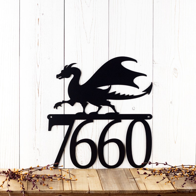 4 digit metal house number plaque with a dragon silhouette, in matte black powder coat. Placed against a white wood wall.