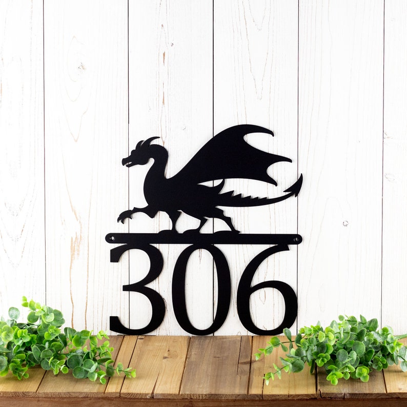 3 digit metal house number sign with a dragon silhouette, in matte black powder coat. Placed against a white wood wall.