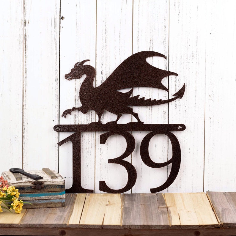 3 digit metal house number sign with a dragon silhouette, in copper vein powder coat. Placed against a white wood wall.