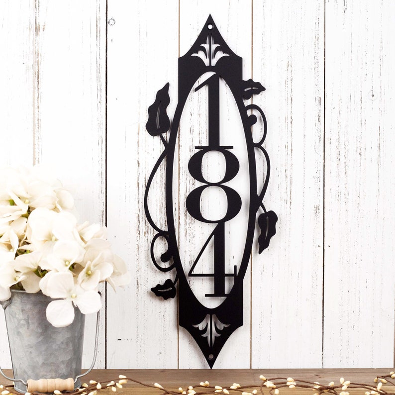 3 digit vertical metal house number sign with vines and fleur de lis, in matte black powder coat. Placed against a white wood wall.