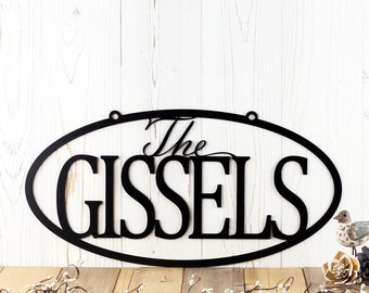 May include: A black metal oval sign featuring the word The in script font above the word GISSELS in a bold serif font. The sign has two circular mounting holes at the top and is displayed against a white vertical wood plank background.